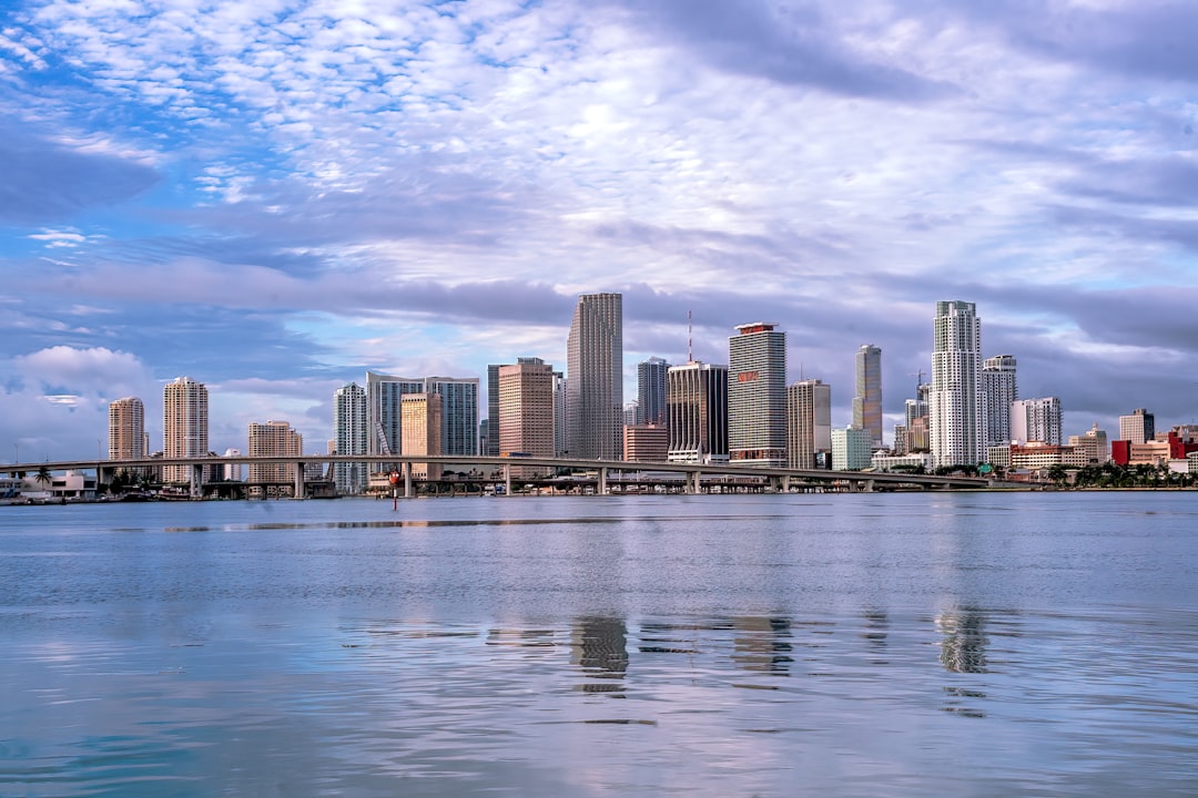 Miami downtown skyline reflected in calm bay waters under a cloudy sky.