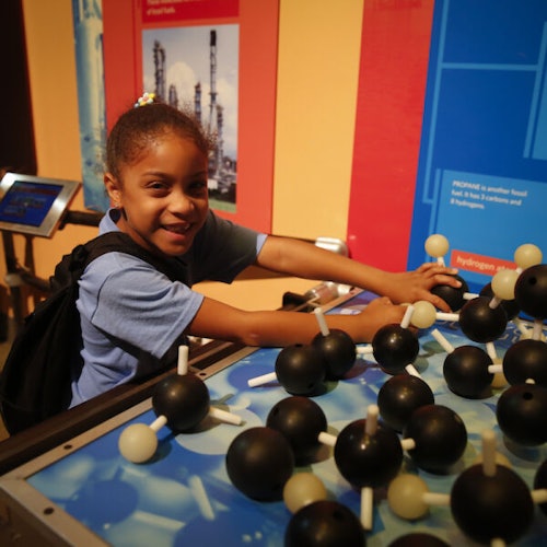 Una joven sonríe mientras explora una exhibición de ciencia con modelos moleculares, vistiendo una camisa azul y una mochila.