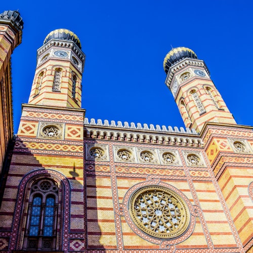 Facciata di edificio ornato con mattoni a strisce, cupole gemelle, finestre intricate e un grande rosone contro un cielo azzurro limpido.