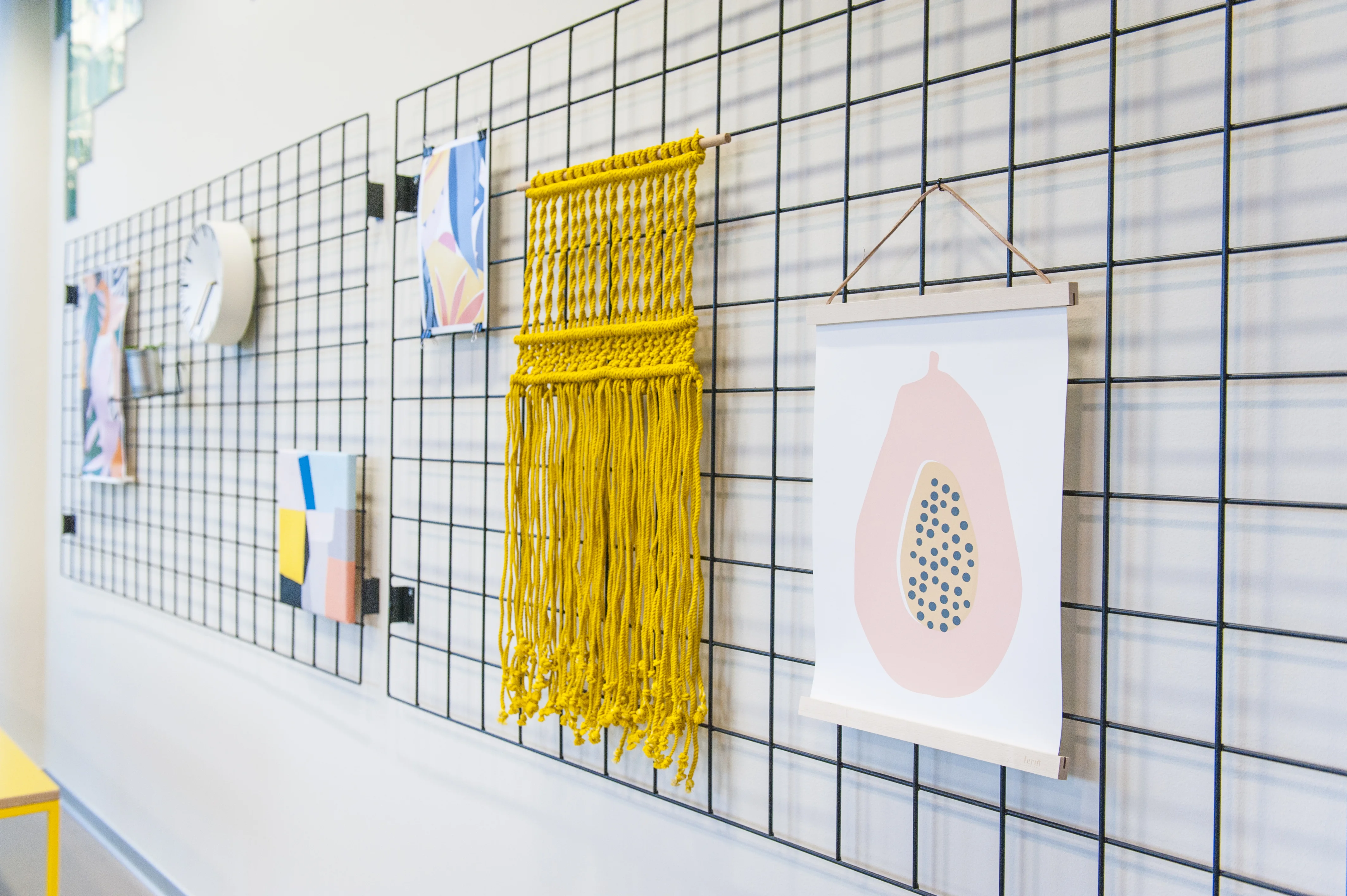 Wire grid wall panels at KANTINE KOMTUR holding a white clock, abstract prints, a papaya poster and a yellow macramé hanging on a white wall.