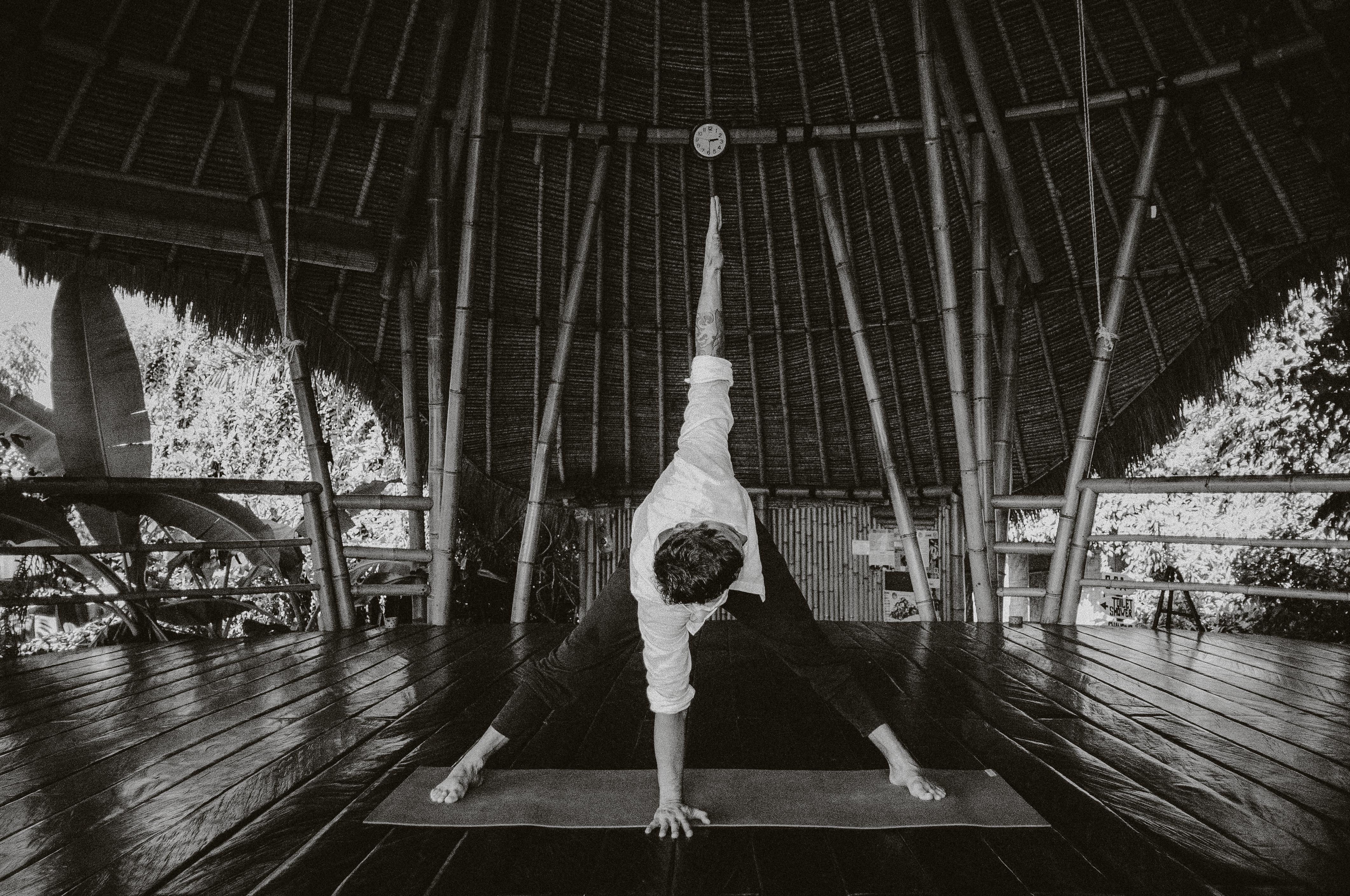 A person practices yoga on a wooden platform in a tropical bamboo pavilion, stretching in a triangle pose under the thatched roof, with lush greenery visible through open sides.