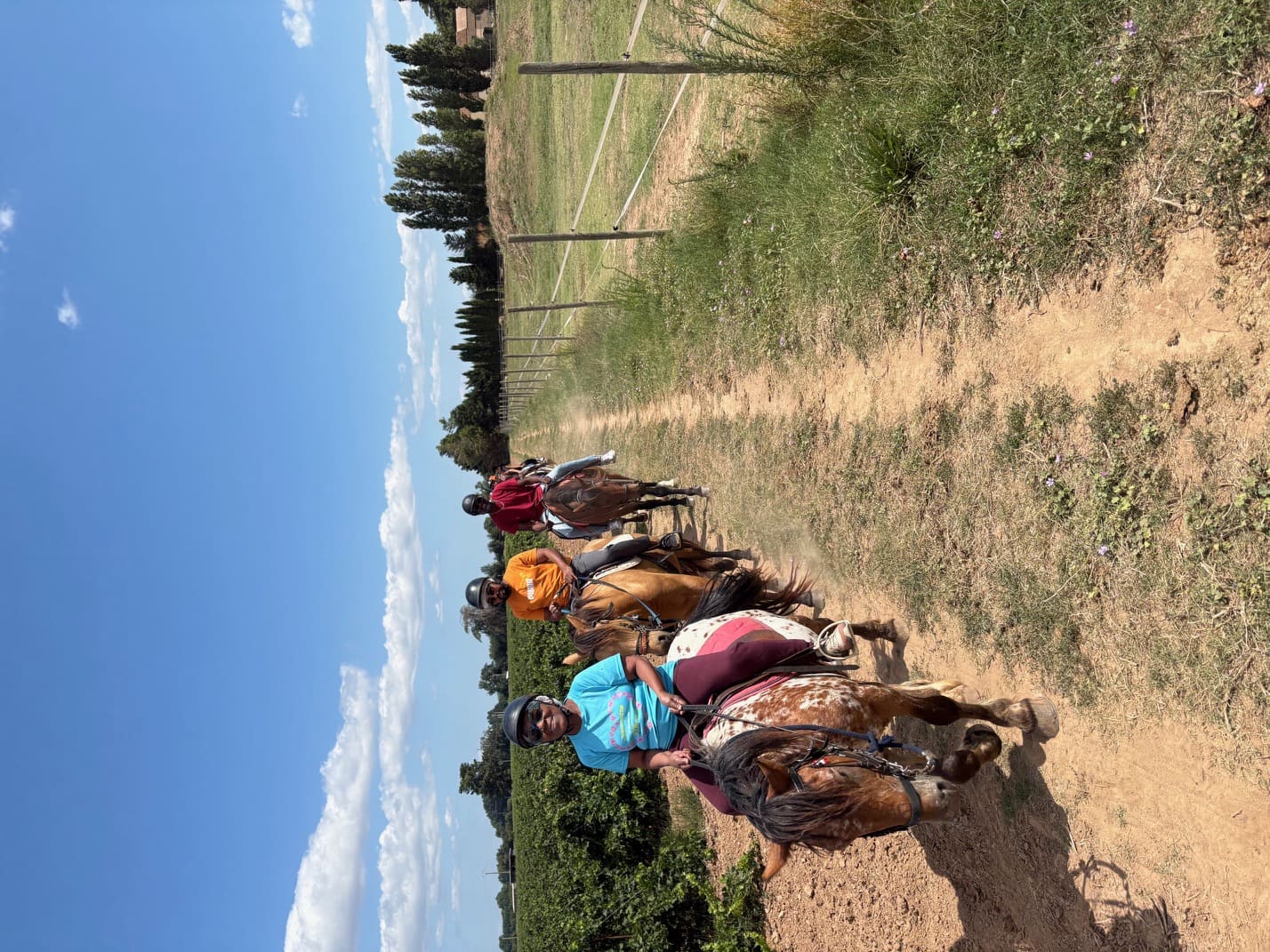Groupe de cavaliers souriants en balade à cheval sur un chemin ensoleillé le long des vignes au Ranch Pegase.