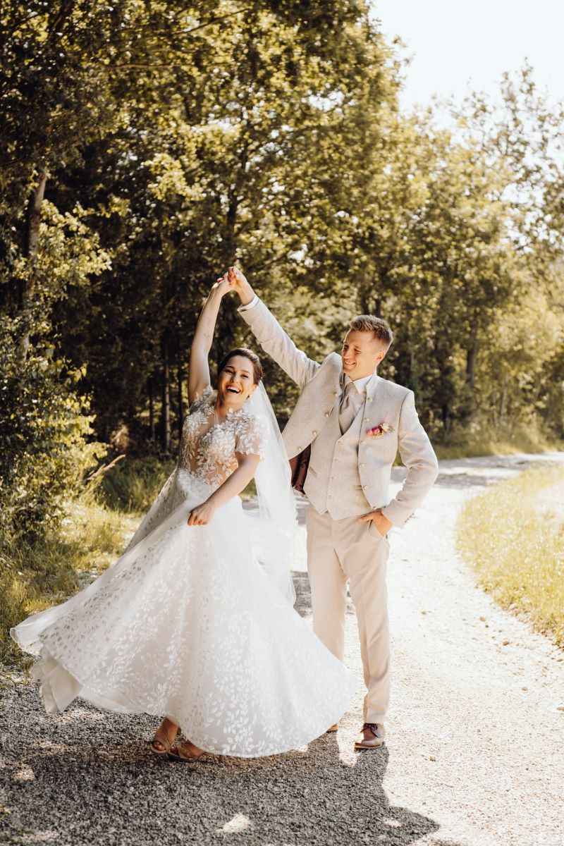 Kundenfeedback Hochzeitspaar für Maria Wiethaler freie Rednerin und Sängerin. Lebendiges Brautpaar in der Natur: Die Braut trägt ein romantisches Spitzen-Hochzeitskleid mit floralen Details und schwingt lachend ihr Kleid, während der Bräutigam in einem stilvollen, hellbeigen Trachtenanzug mit Weste und Krawatte ihre Hand hält. Beide strahlen Freude und Leichtigkeit aus, während sie auf einem idyllischen Waldweg im Sonnenlicht tanzen. Authentische Hochzeitsfotografie mit natürlicher Eleganz und emotionalem Charme.