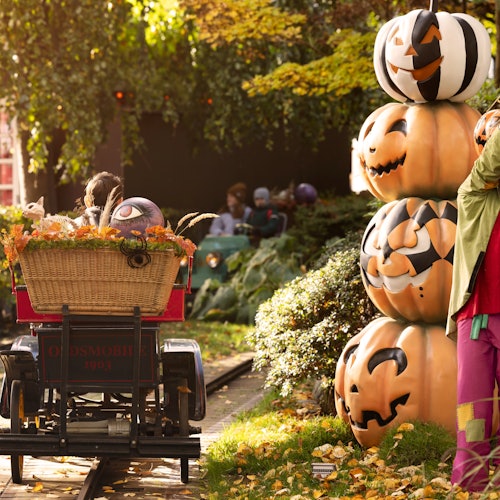 Folk kører i en vogn med vintagetema med en kurv af efterårsdekorationer, mens de passerer en bunke udskårne græskar i en park-lignende omgivelser.