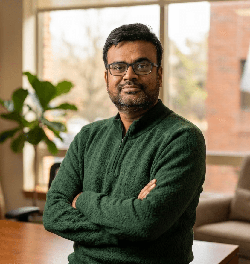 A smiling person with curly hair sits at a desk, looking at a laptop against a dark background.