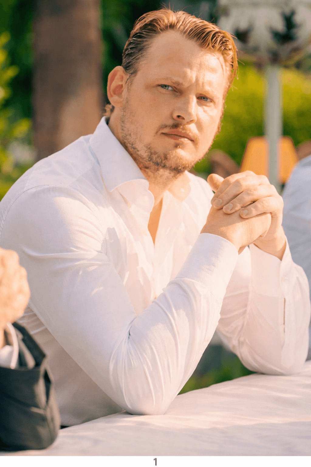 A young man with light brown hair is wearing a black suit jacket and white shirt, standing in a softly lit office setting, conveying professionalism.
