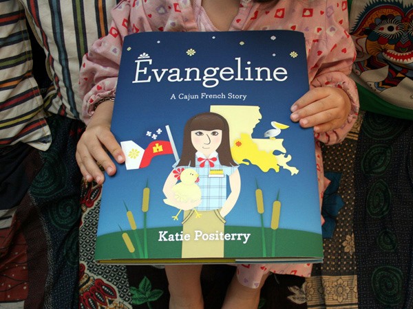 A child is holding a book titled "Evangeline: A Cajun French Story" by Katie Positerry. The cover features a cartoon illustration of a girl in a plaid shirt and khaki skirt standing in a grassy field with a chicken and a flag. The background includes a stylized map of Maine and a bird. The book is held against a colorful, patterned fabric.