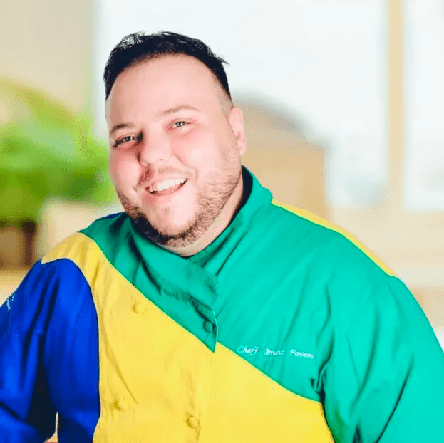 A young person with curly hair smiles at the camera while sitting in front of a laptop against a dark background.