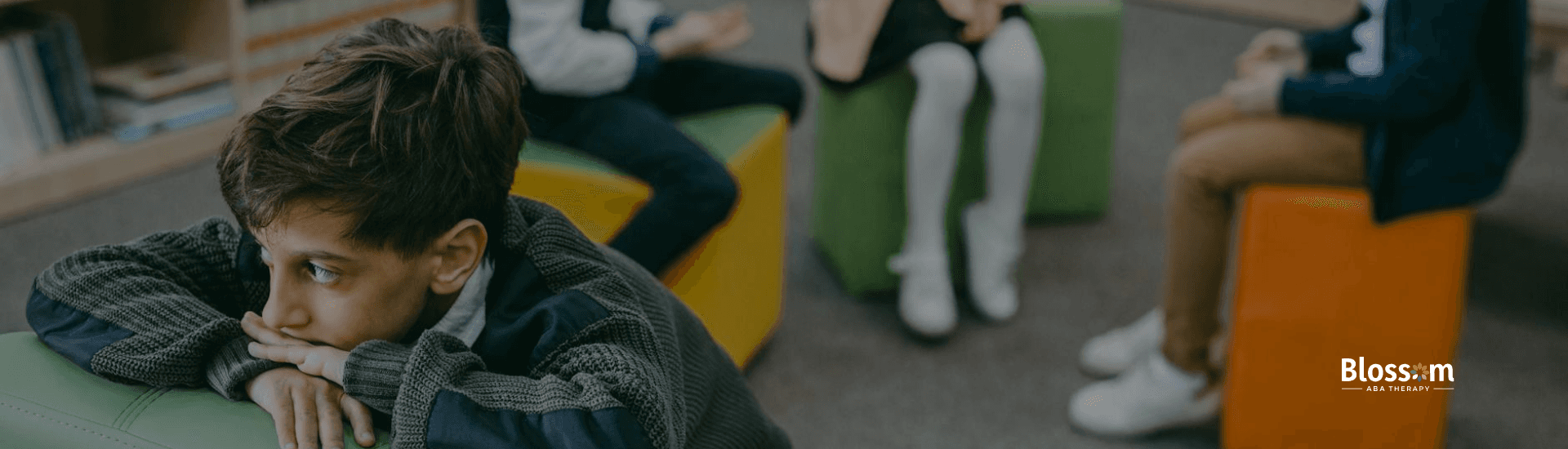 An autistic boy sits apart from a group of children in a library, appearing sad and withdrawn in GA.