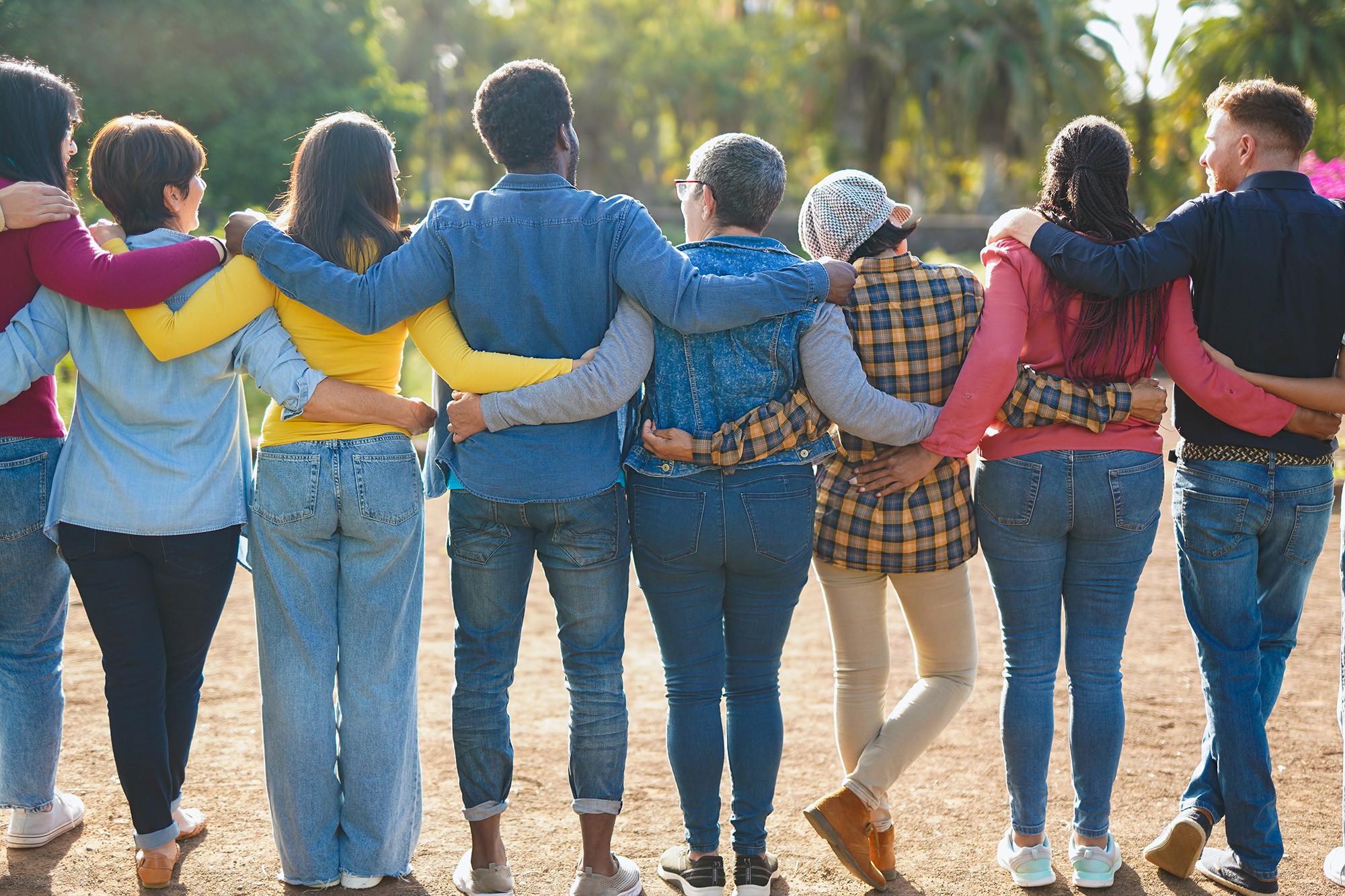 Un grupo diverso de nueve personas de pie con los brazos entrelazados, vistas desde la espalda, representando unidad y amistad