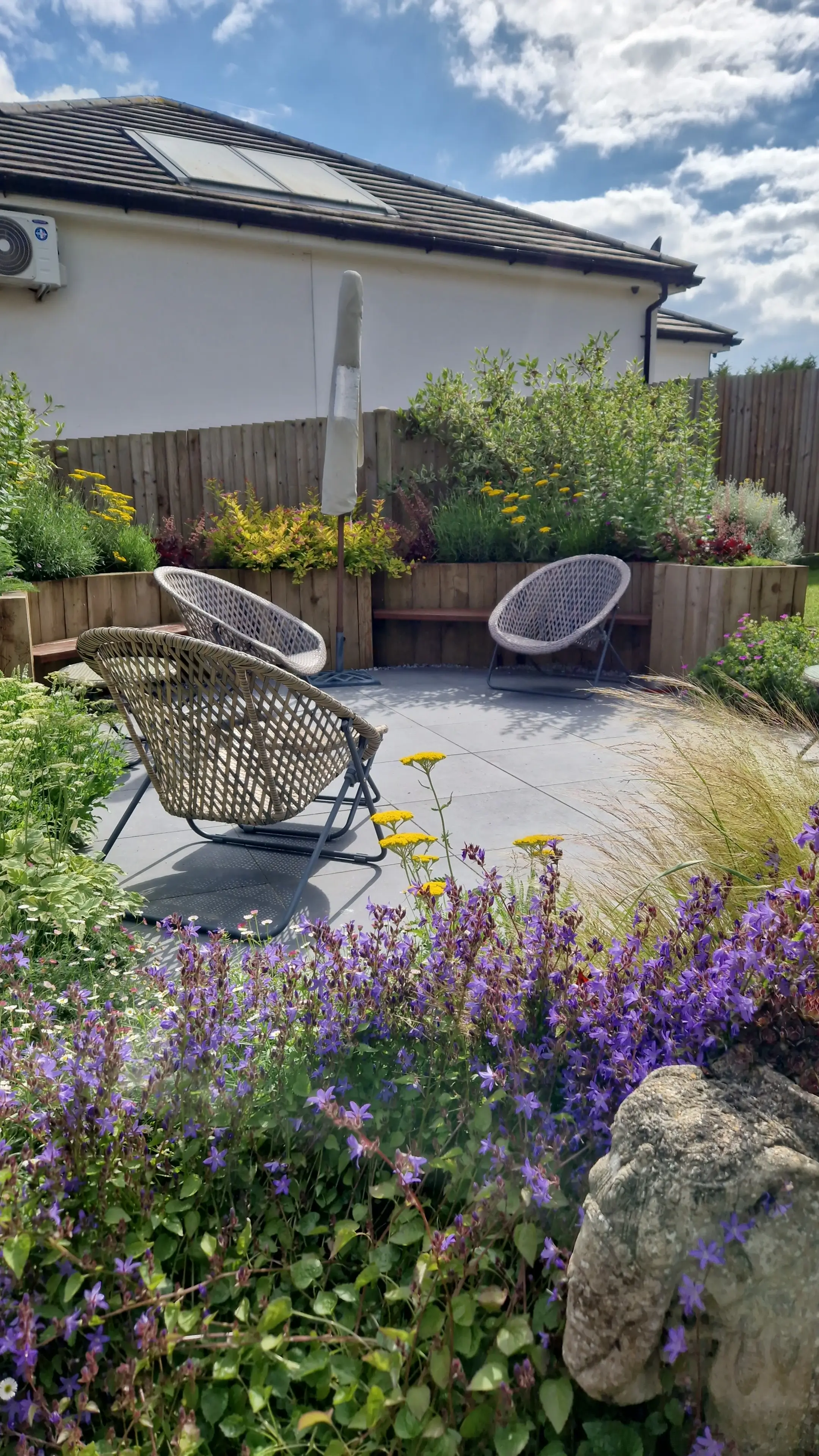 A vibrant garden featuring colorful flowers, rocks, and a pathway leading to a house under a blue sky.