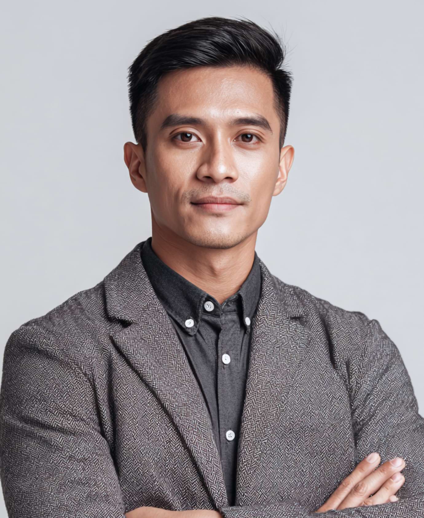 A confident man with short hair stands with arms crossed. He wears a gray blazer over a black button-up shirt. The background is plain gray.