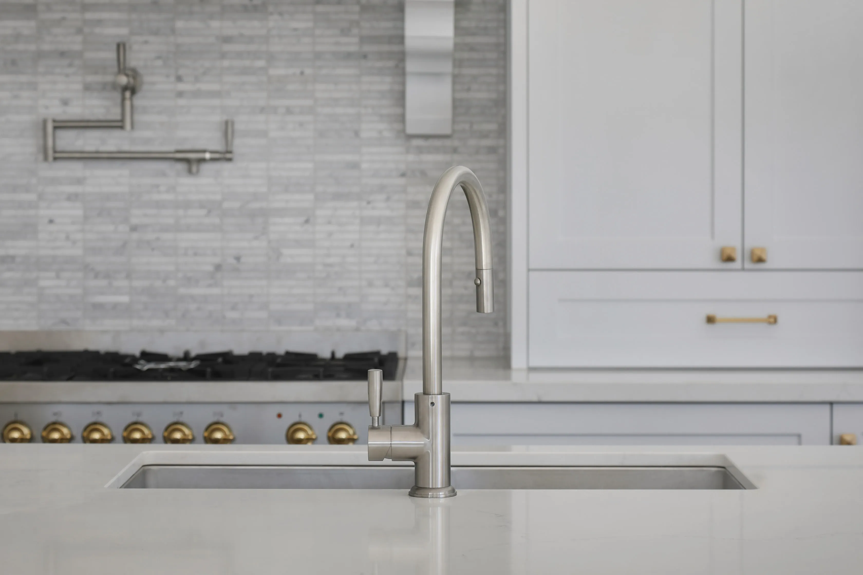 Kitchen detail with a brushed-nickel gooseneck tap and pot filler over a stainless undermount sink in a white benchtop, with a marble mosaic splashback and a brass-handled gas range behind