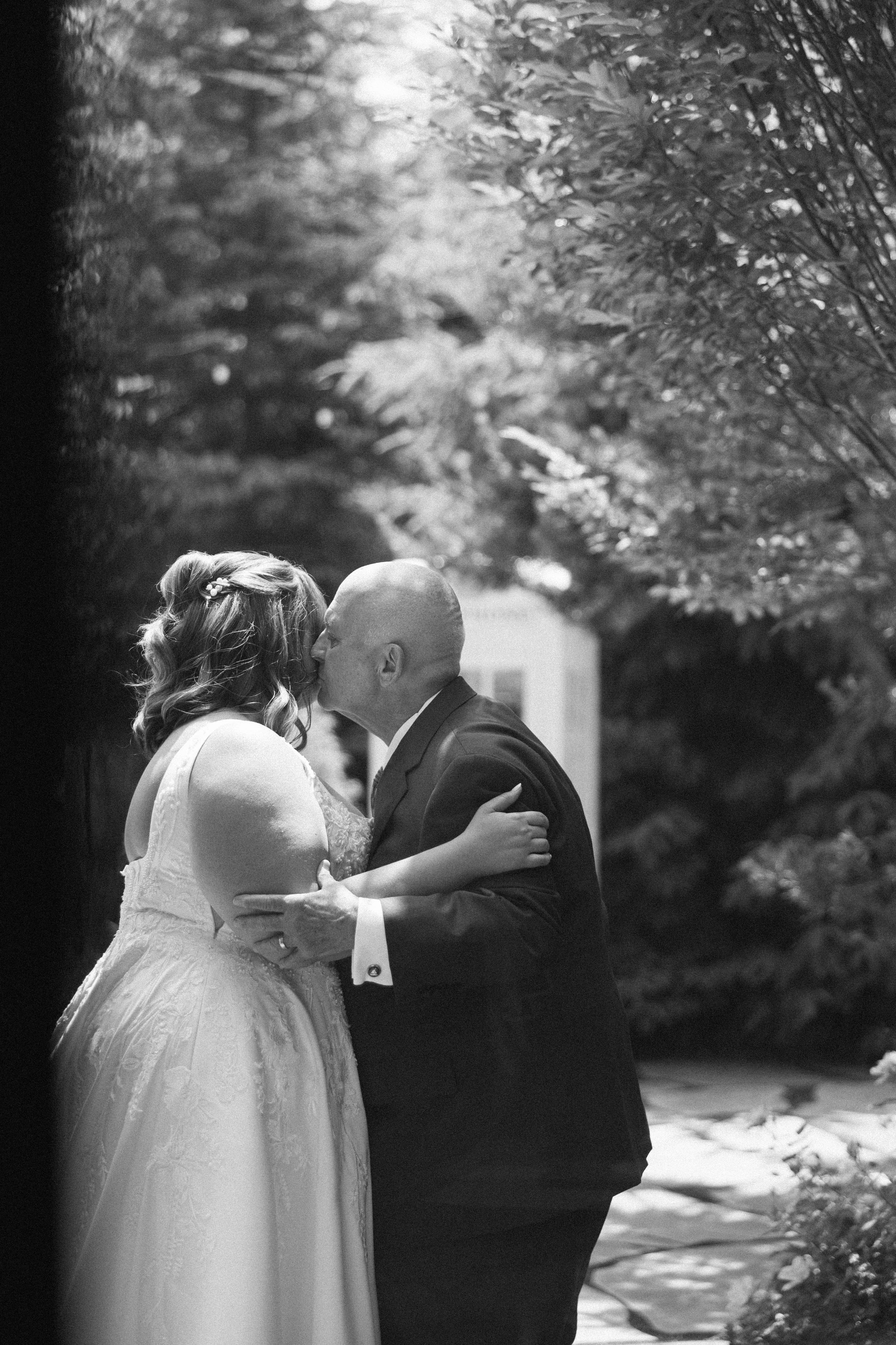 A black-and-white photo shows a man kissing a woman on the cheek. Both are dressed in formal attire, and they are surrounded by trees.