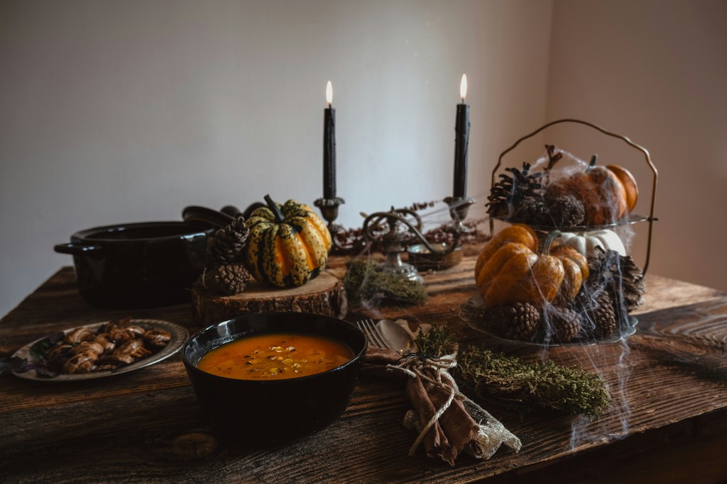 A cozy autumn table setting features a bowl of pumpkin soup, a variety of pumpkins, pine cones, dried herbs, and two lit black candles, creating a warm and festive atmosphere perfect for a fall or Halloween gathering.