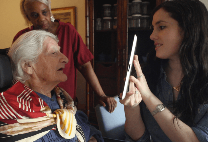 An older woman with white hair sits in a chair wearing a red, white, and tan patterned scarf. She looks toward the woman on the right, who is holding a tablet and looking at the older woman. The older woman appears to be speaking or reacting to the tablet. The setting is a room with a wooden cabinet or bookcase in the background and a framed picture on the wall. A second woman in a red shirt is standing behind the older woman, partially visible.