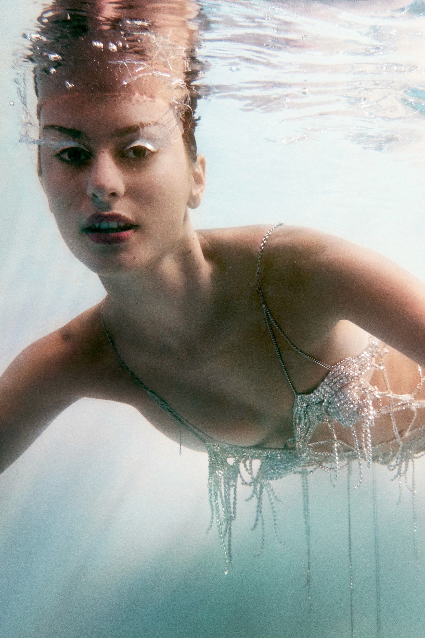 Underwater fashion photography of a model lying face-down on the water's surface, wearing a dress made from strings of sparkling jewels that hang from her body.
