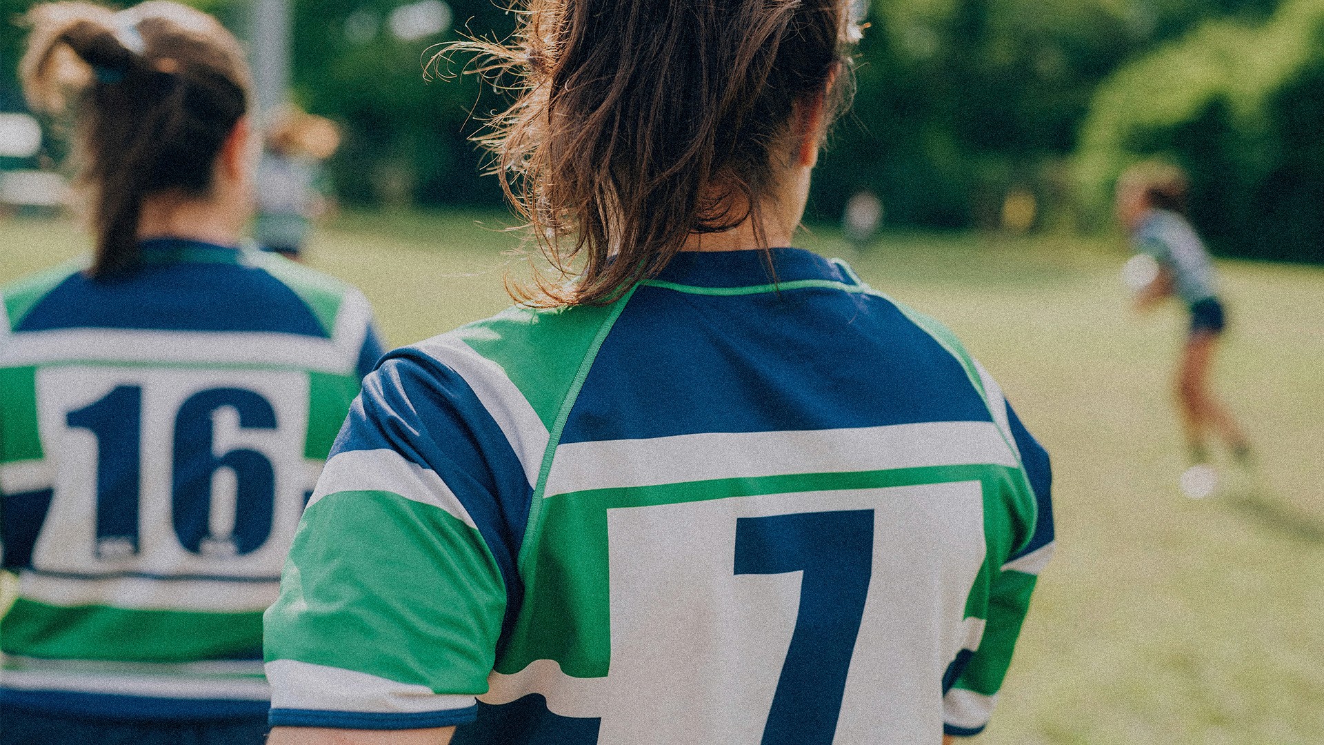 3 women in frame on a field wearing blue, green, and white rugby jerseys. One jersey says 16, the other says 7, and the third is out of focus and far away.