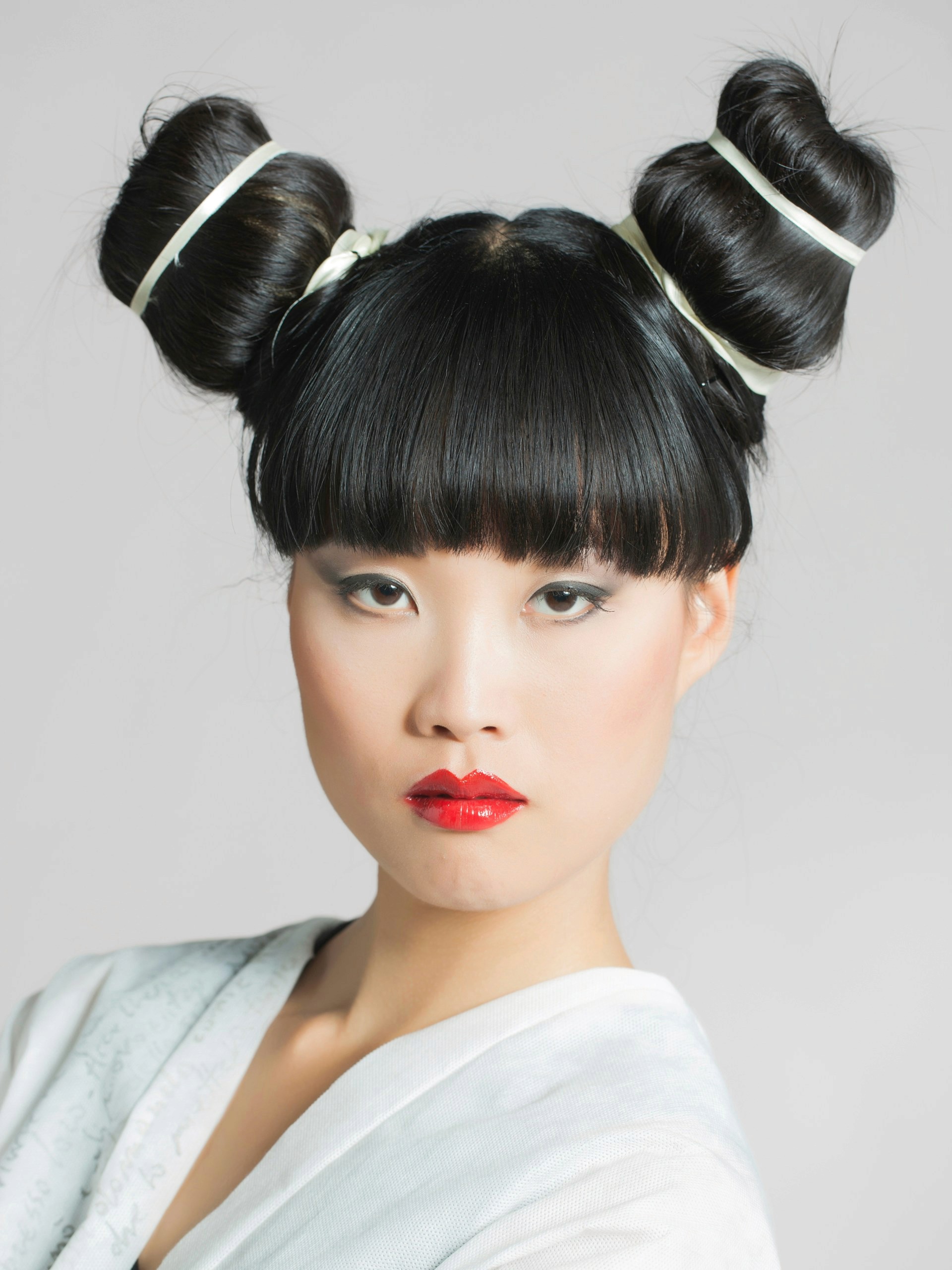 A high-fashion portrait of an East Asian woman with blunt bangs and her hair styled in two large, structured buns, wearing bright red lipstick.