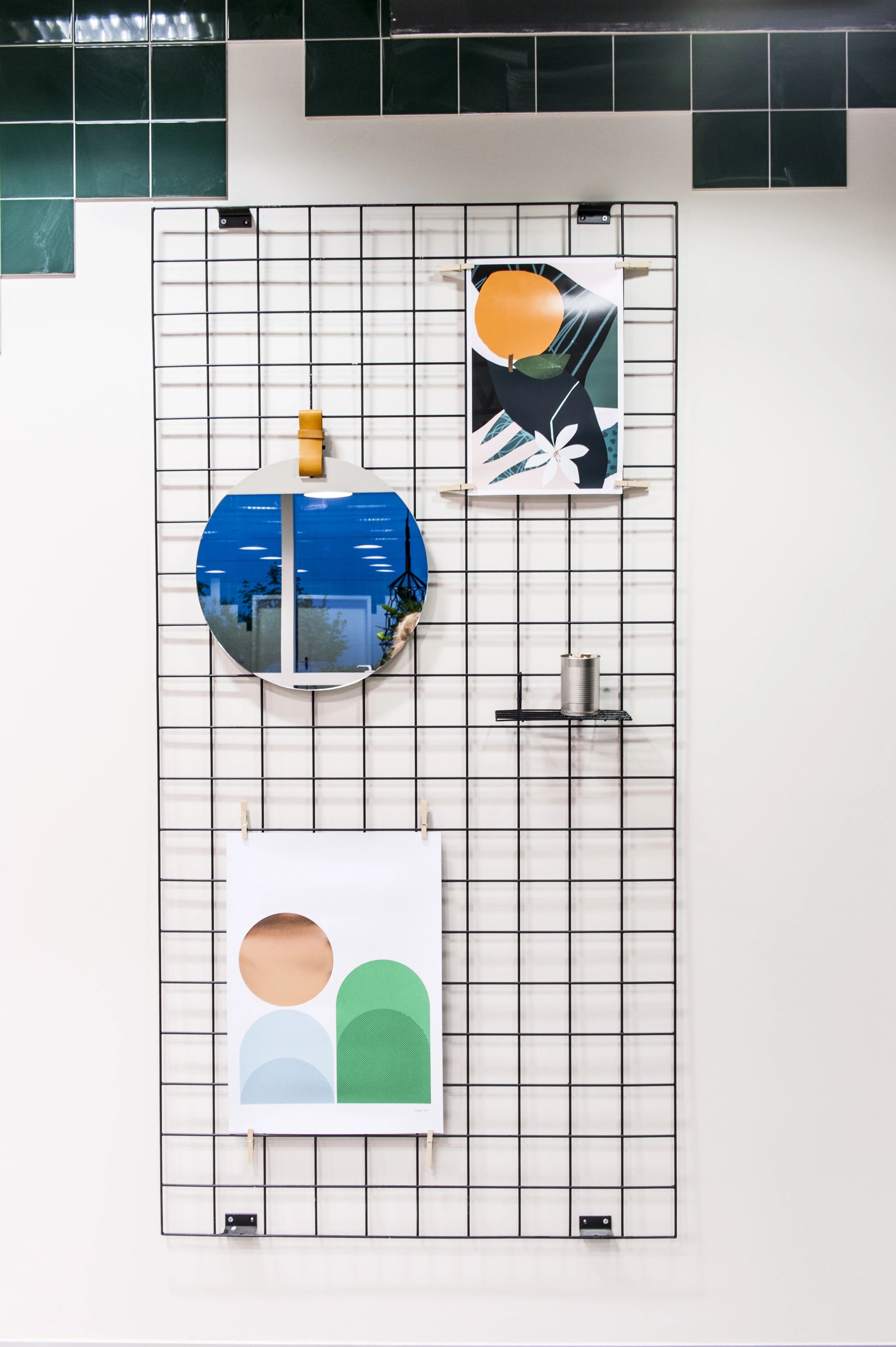 Vertical wire grid panel at KANTINE KOMTUR displaying a round mirror, graphic art prints and a small metal shelf beneath green pixel-pattern tiles.