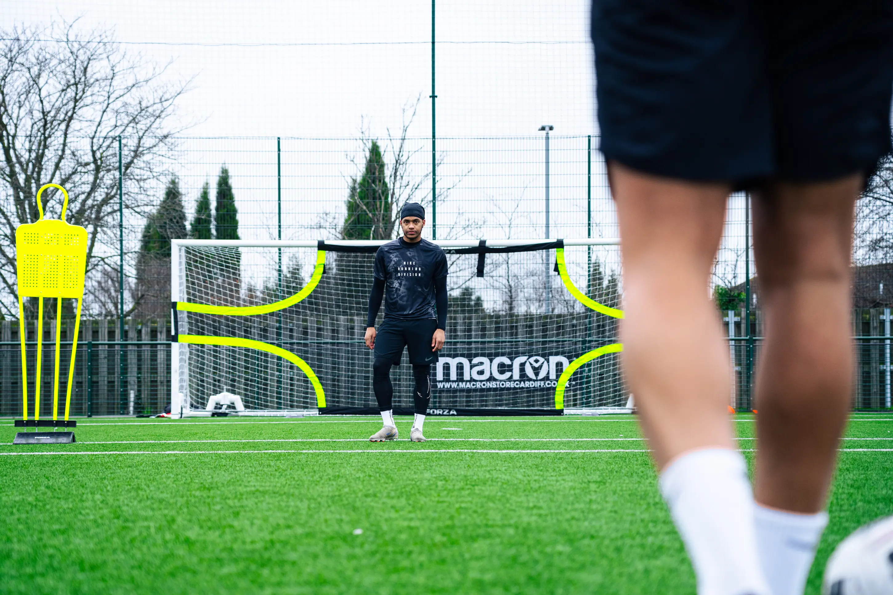 A football player wearing a dark outfit stands in focus in front of a large soccer goal net on a green field.