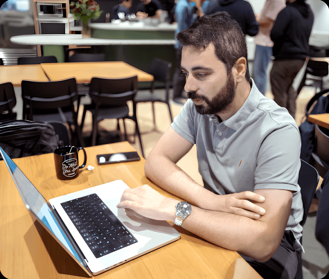a man sitting at a table, focused on his laptop. He has a beard and is wearing a polo shirt. A coffee mug is placed nearby, and he appears to be deep in thought or engaged in work.