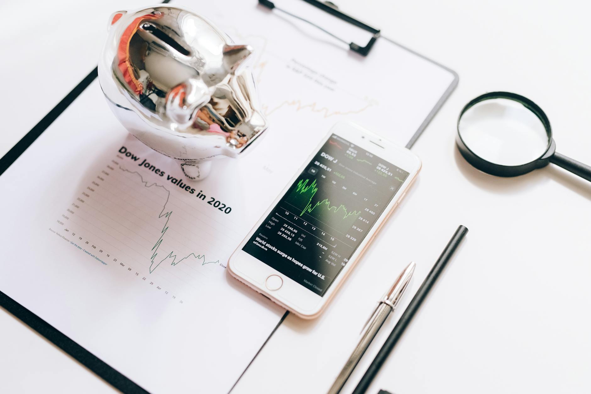 Silver piggy bank and smartphone displaying stock market charts on a desk with financial reports and a magnifying glass