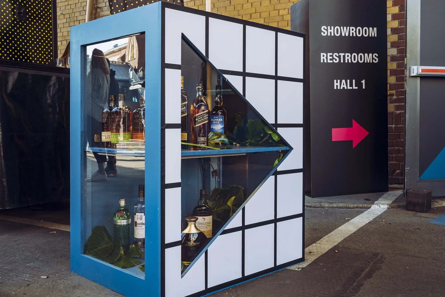 Blue display cabinet with black grid front and triangular glass cutout holding whisky and gin bottles outside DIAGEO BCB19, next to a showroom sign.