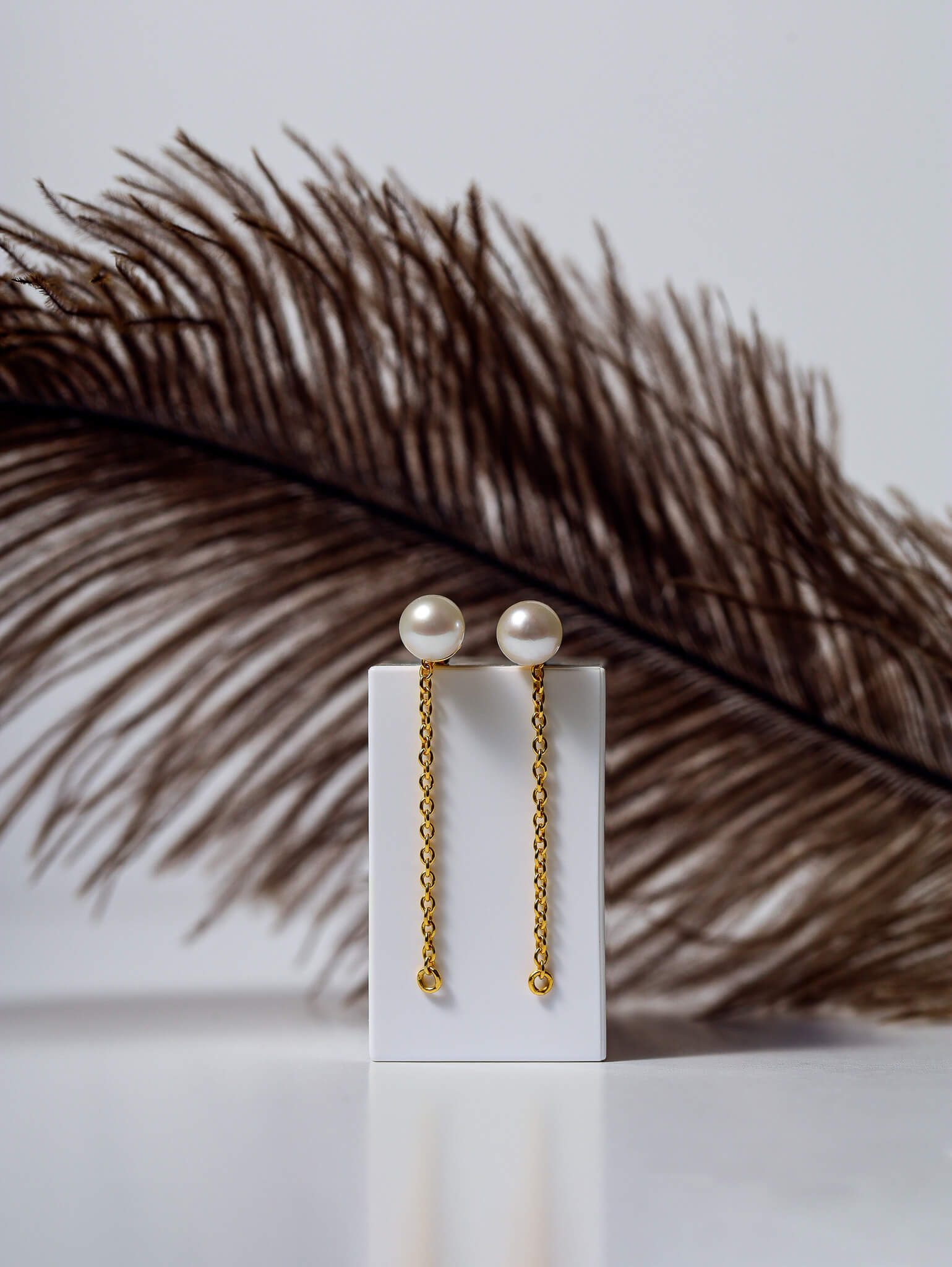 Minimal product photography of pearl drop earrings with gold chain detail and feather backdrop.