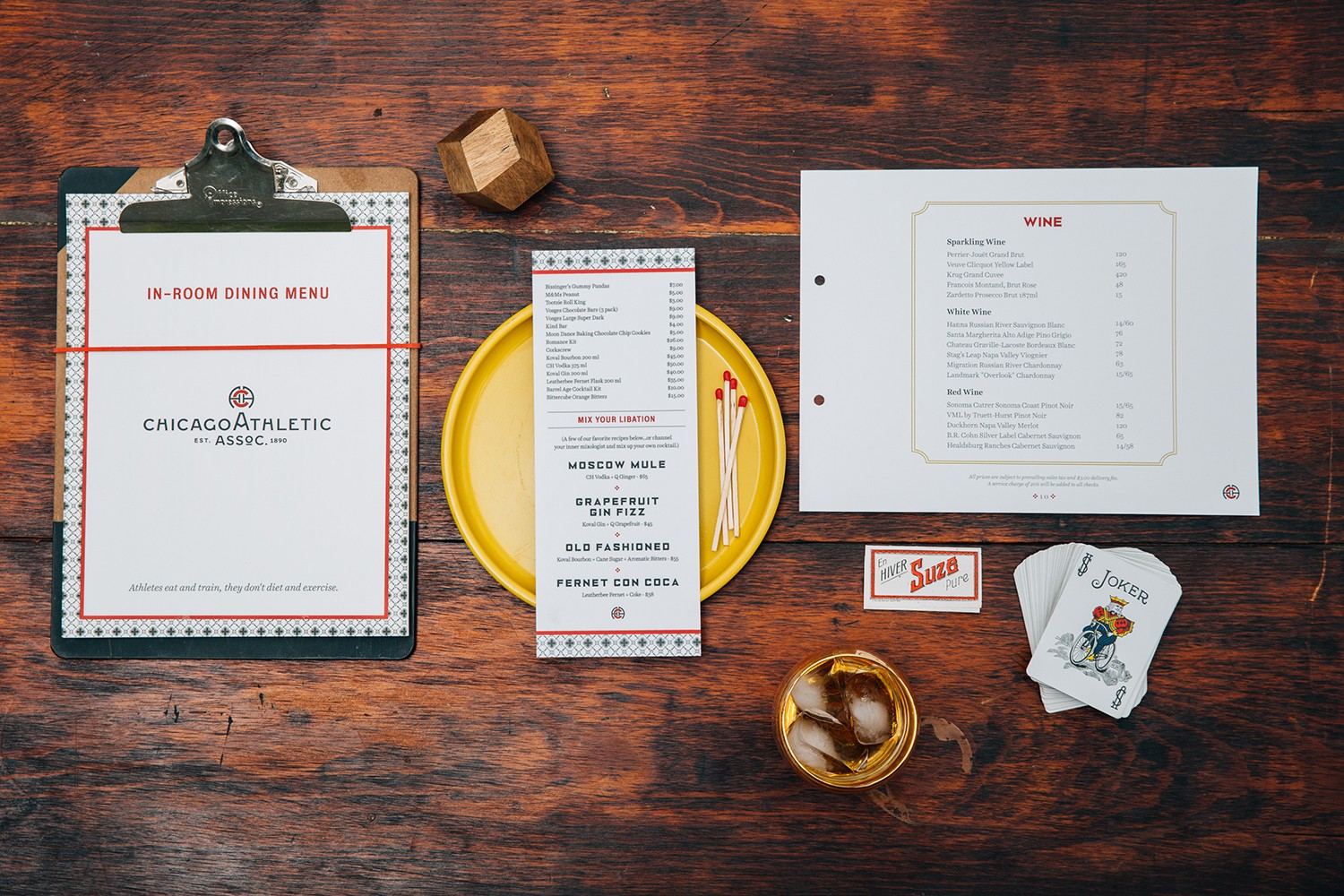 Menus styled with a whiskey cocktail, yellow tray, matches, vintage rolling papers, and a deck of cards showing the Joker.