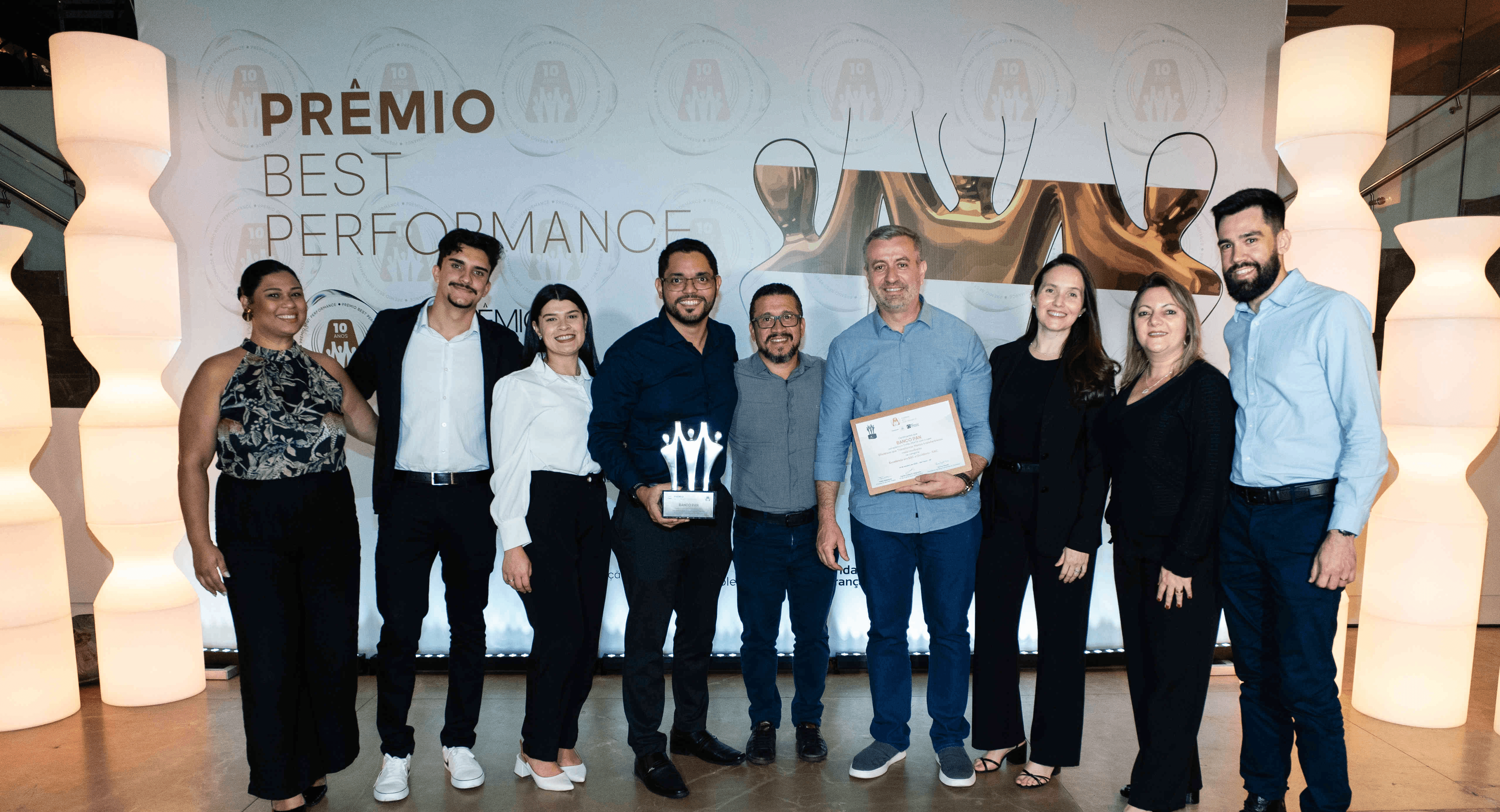 Executivos do BANCO PAN posando com troféu prata e certificado no palco do Prêmio Best Performance 2025