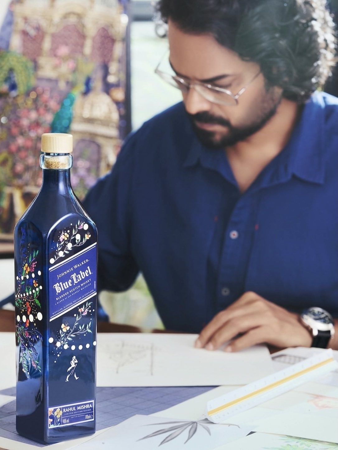 A close-up of a limited edition Johnnie Walker Blue Label bottle designed by Rahul Mishra, with vibrant floral and animal motifs, placed on a table beside a designer sketchbook and tools, featuring a male designer in the background.