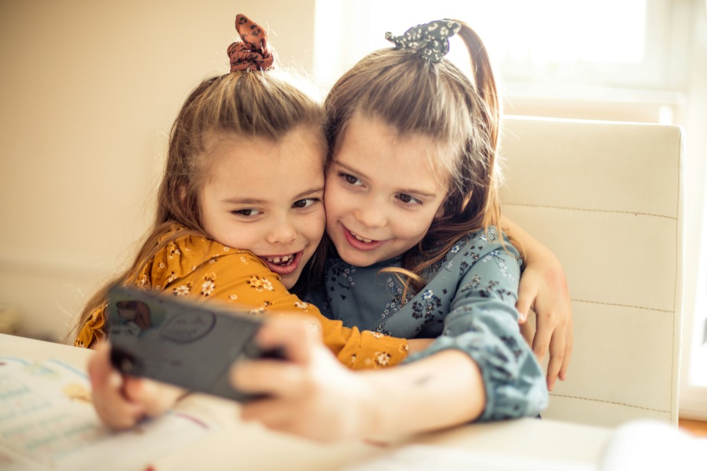 Two young girls, dressed in colorful outfits with hair tied up in matching scrunchies, cuddle and smile as they take a selfie together. The scene captures their close bond and joy, highlighting the innocence and happiness of childhood moments.