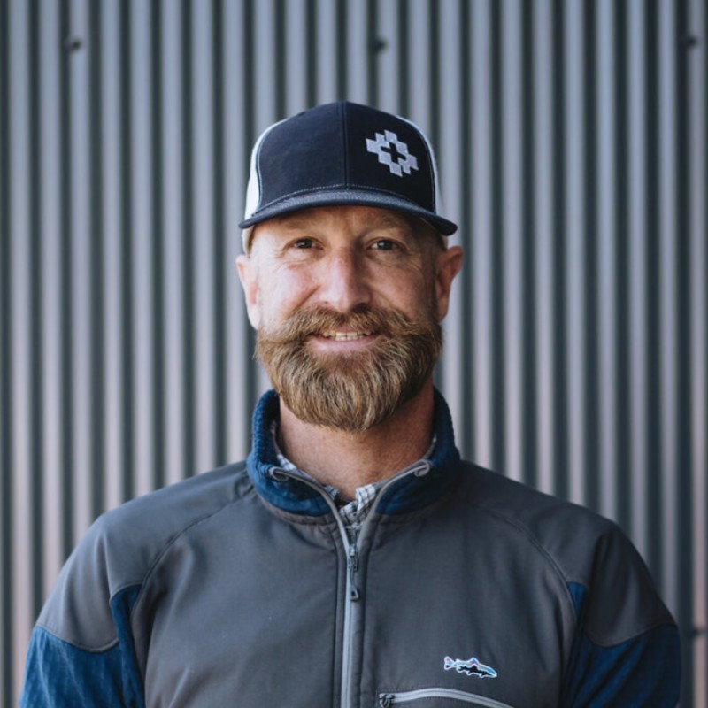 A portrait of a white mnan smiling with blond hair, a blue baseball cap, a thick blond beard, and a sport zip-up jacket