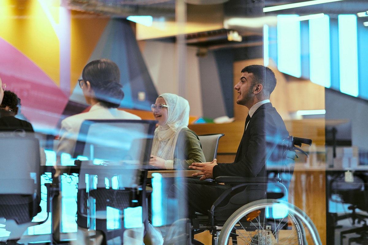 A diverse team of five colleagues smile while gathered around a glass wall covered with colorful sticky notes in pink, yellow, and green. The group appears engaged in collaborative brainstorming or planning, with one woman in the foreground actively placing or organizing notes. The casual, energetic atmosphere suggests creative problem-solving and inclusive teamwork.