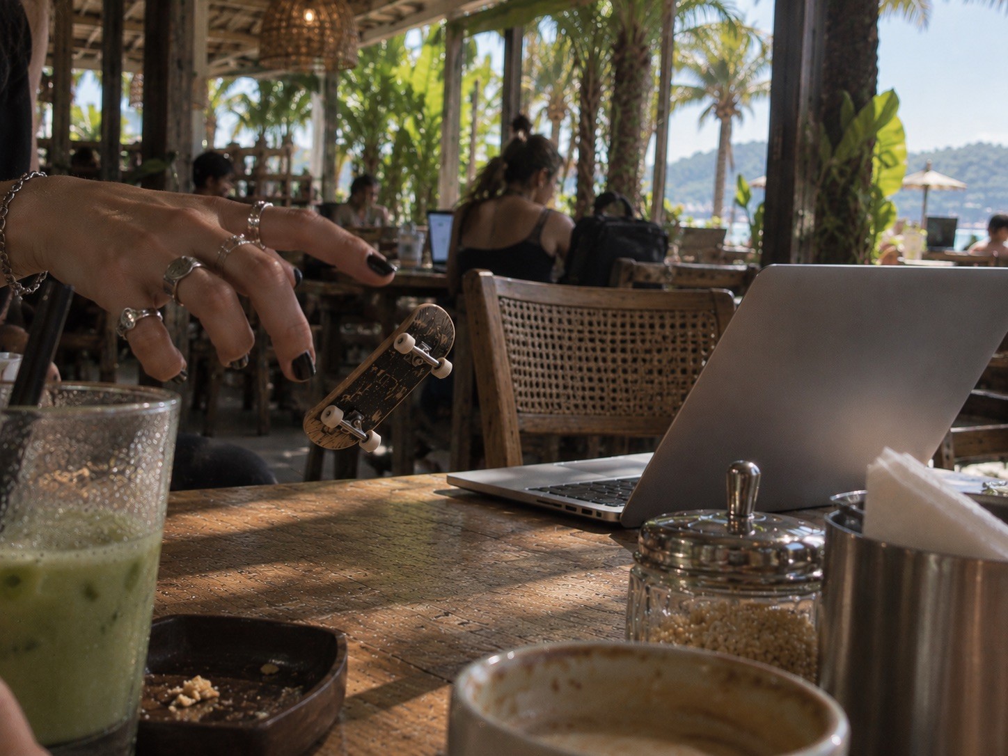 A hand with black nail polish and silver rings flips a miniature skateboard beside a laptop at a sunlit tropical café.