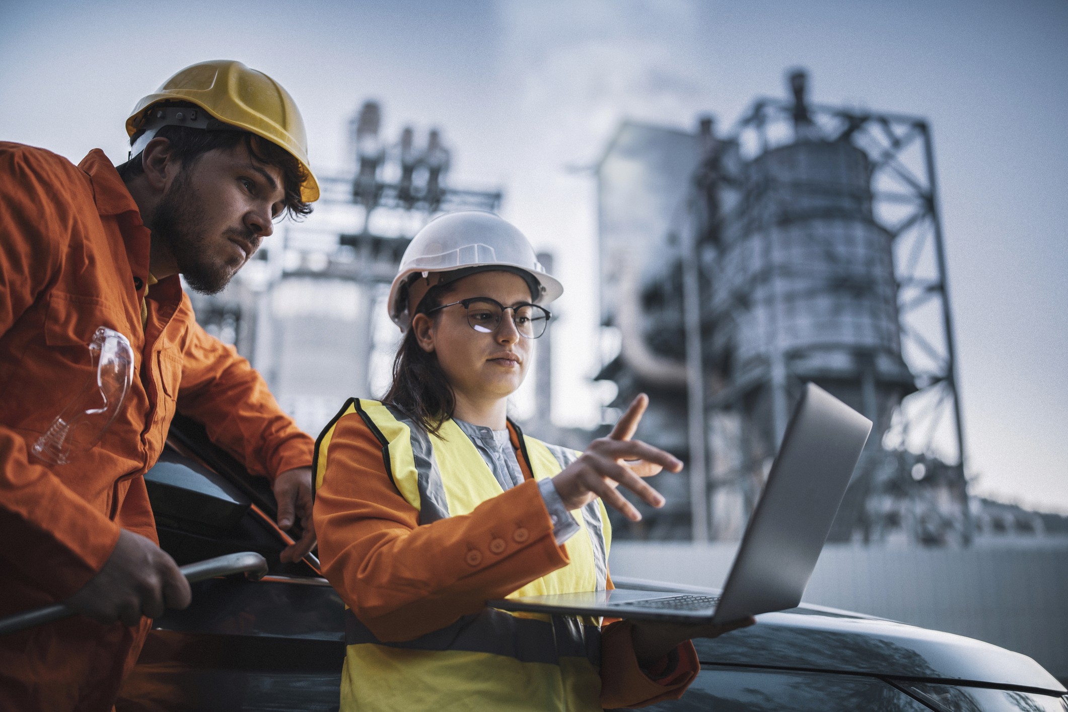 Two workers in safety gear discuss a laptop in an industrial setting with machinery in the background.