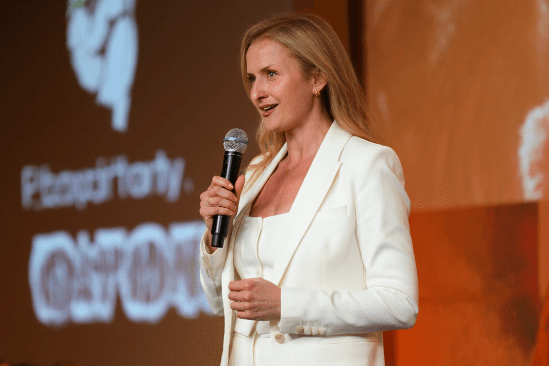 Two women are standing side by side against a beige wall, smiling confidently, one wearing a green shirt and the other in a dark blazer, with soft lighting casting gentle shadows.