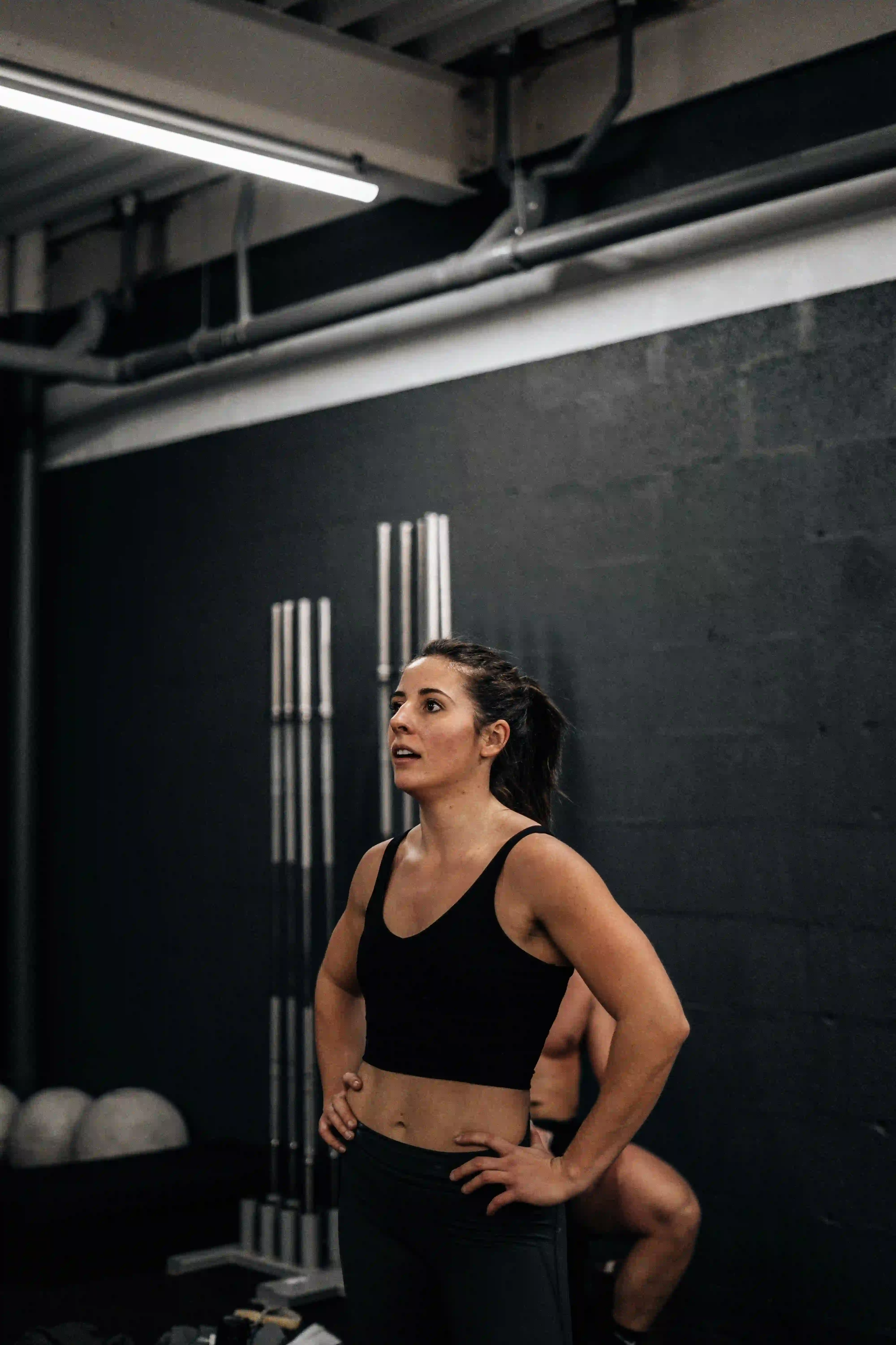 Female athlete standing in a dark gym with hands on hips, catching her breath after a workout.