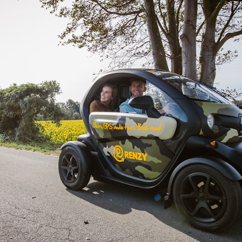 两个人在乡村道路上，一辆标有“RENZY”的小型黑色迷彩电动汽车里微笑，周围是树木和黄色花朵。