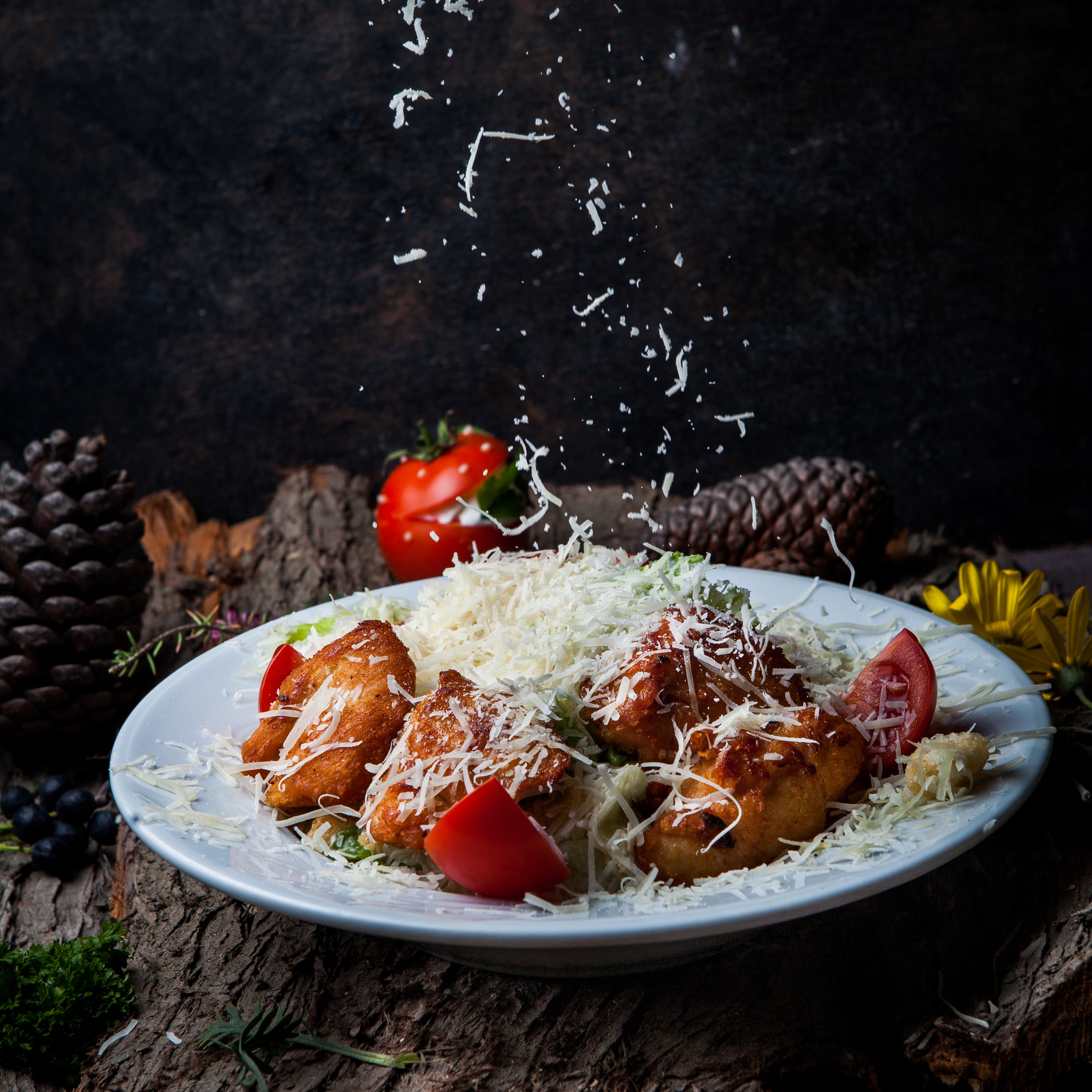 Four pieces of fried chicken or battered fish on a bed of salad greens, being heavily topped with shredded white cheese that is raining down onto the dish, set against a dark, rustic background of wood and pine cones.