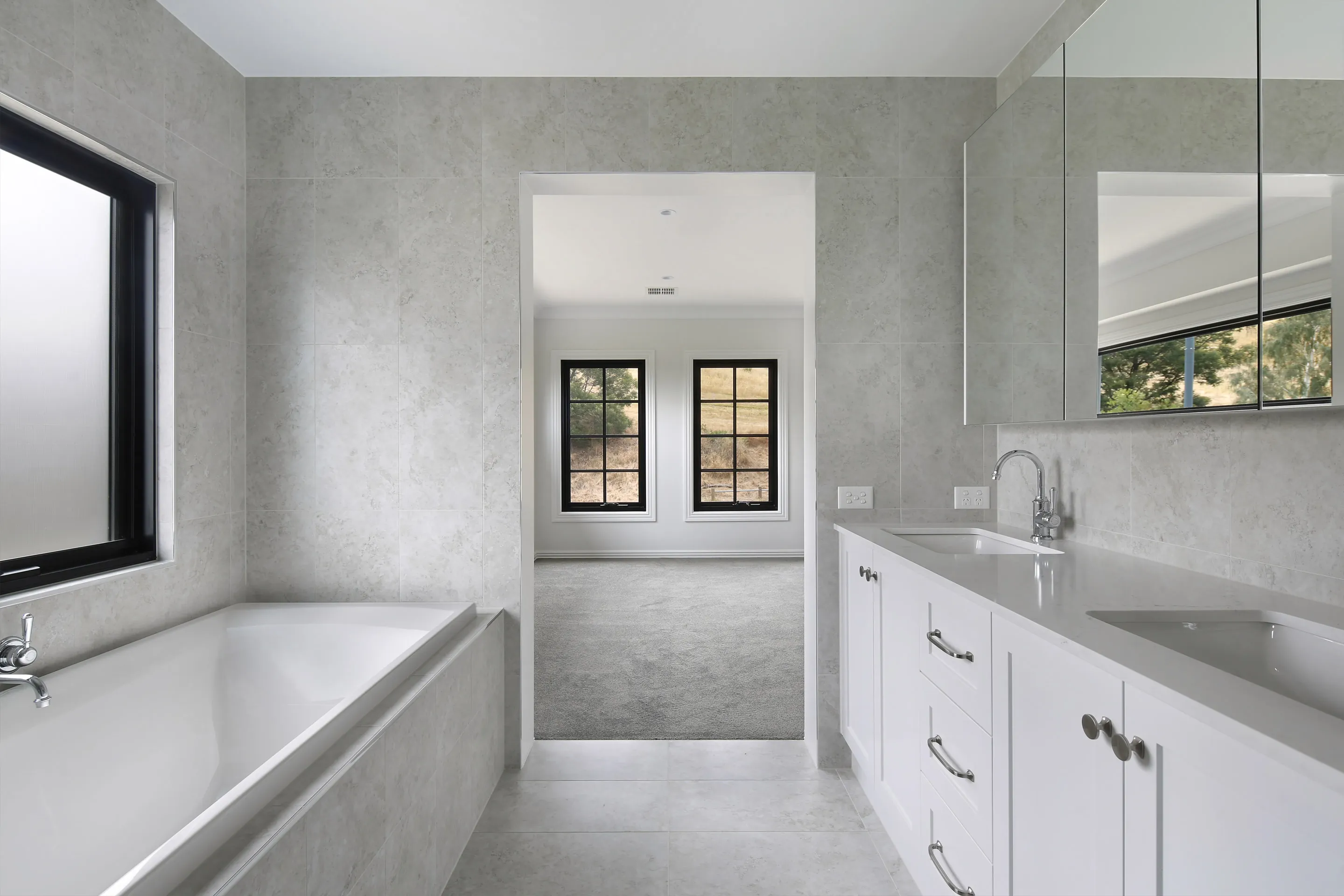 Ensuite with a built-in bath, white shaker double vanity with nickel tapware and mirrored cabinets, opening to a carpeted bedroom with black-framed windows and a rural view