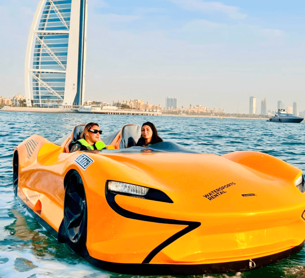 Two women riding a jet car with Dubai's iconic sail-shaped building in the background.