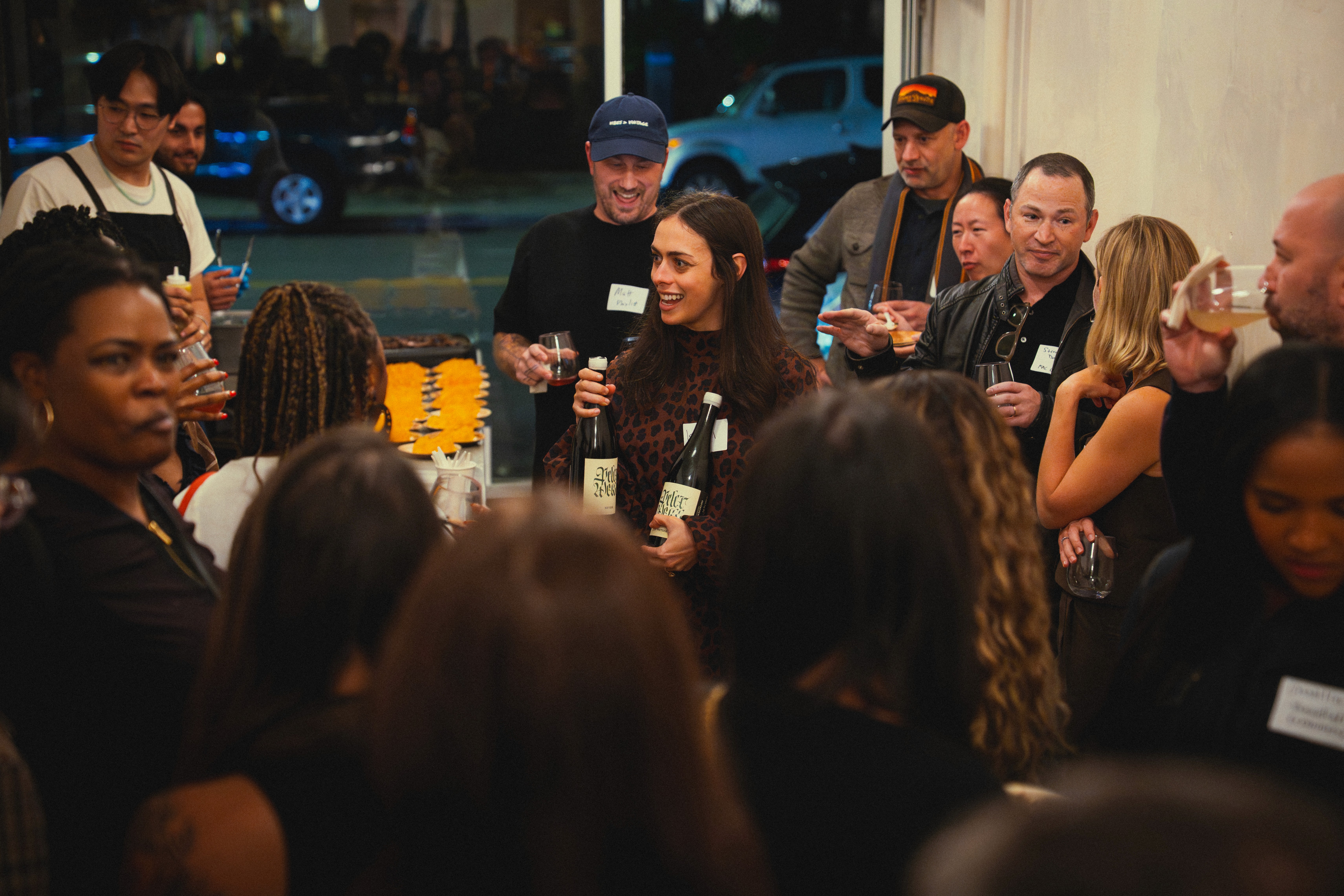 A woman holding two bottles of wine in a room full of people gathering and talking.