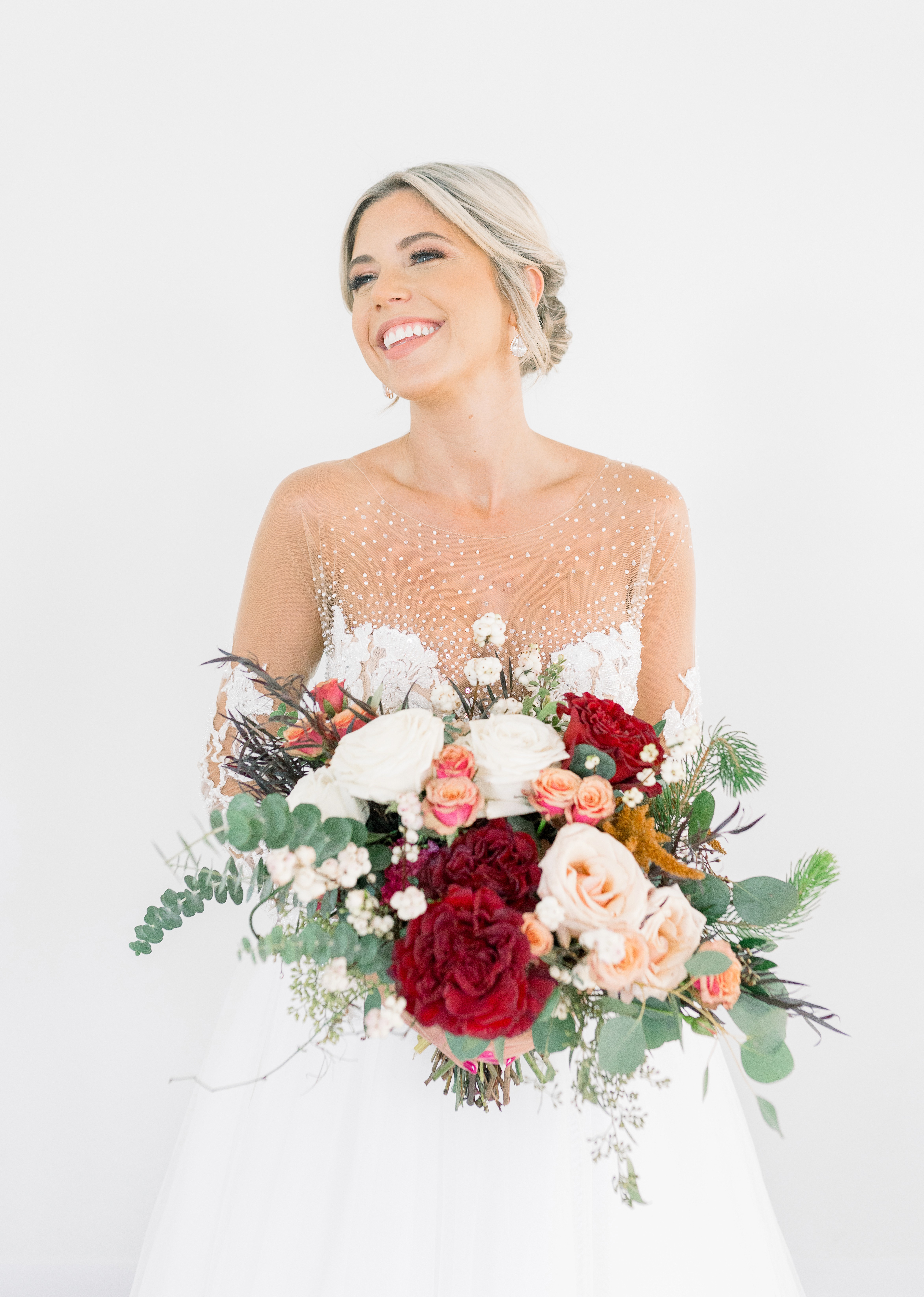 Smiling bride in a sheer pearl-detailed gown holds a large bouquet with red peonies, blush and white roses, eucalyptus, and greenery against a white background.