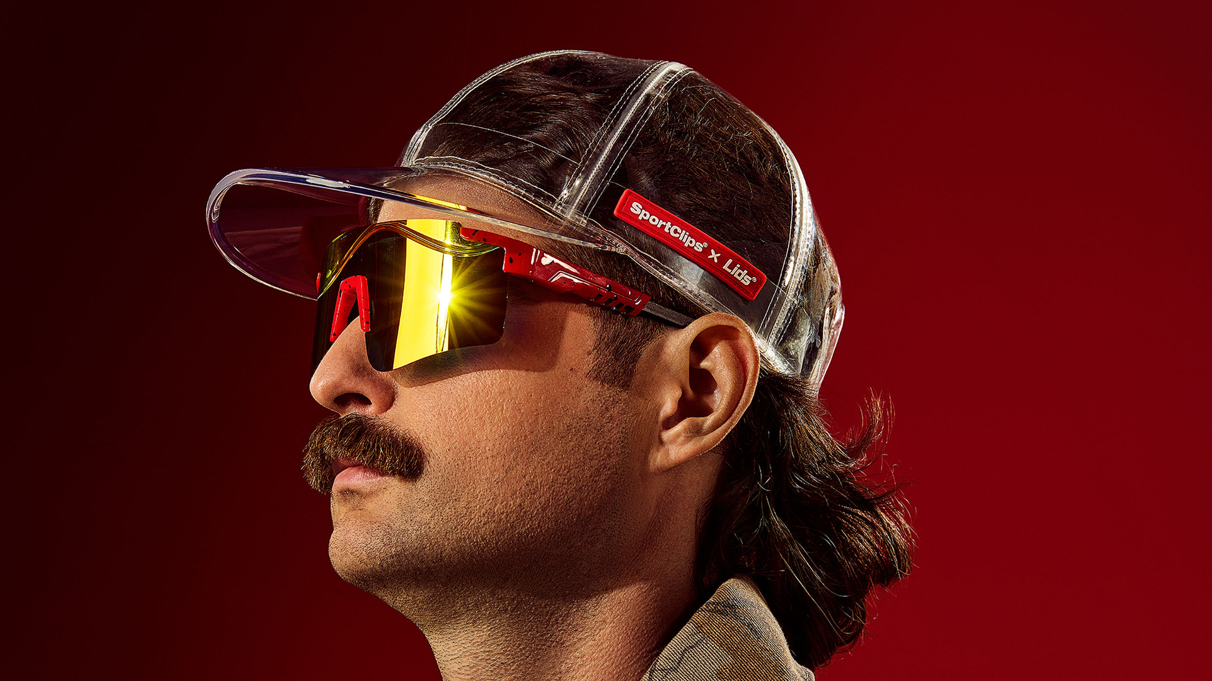 Person wearing a transparent cap against a red background. Only the lower portion of their face is visible, showing a distinctive mustache. The lighting creates a stylized, promotional atmosphere.