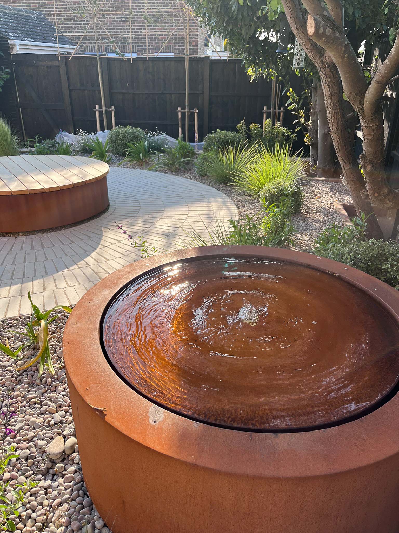 A circular wooden planter sits in a garden, surrounded by green grass and plants under a bright sky.
