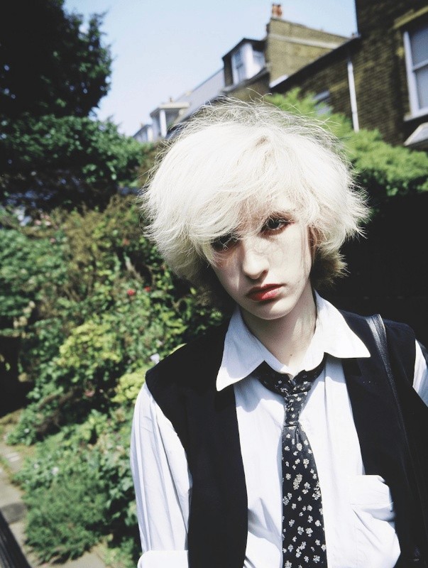 Portrait of a person with white hair and a black vest against a green garden background.