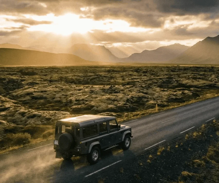 An off-road vehicle driving along a road through green lava fields towards the setting sun, with rays breaking through dramatic clouds.