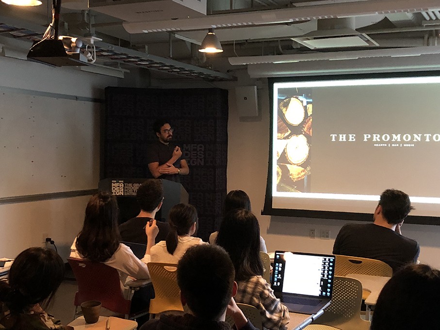 A photo of a man giving a lecture to a class while looking at a projector screen.